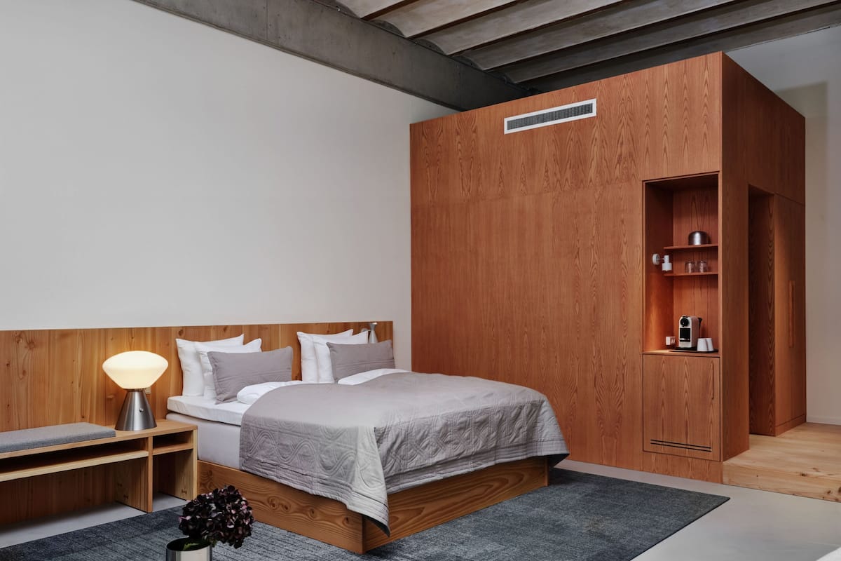 Une chambre moderne avec une tête de lit en bois, un lit avec une literie grise, une table de chevet avec une lampe et une armoire intégrée avec des étagères.