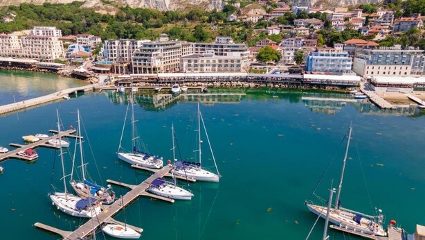 A marina with multiple sailboats docked at piers, surrounded by residential buildings and a hilly landscape in the background.