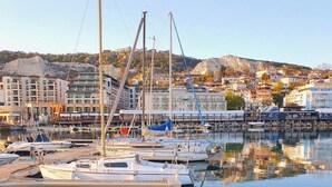 A marina with multiple sailboats docked, a calm water surface reflecting the boats and buildings, and a backdrop of residential buildings and hills.
