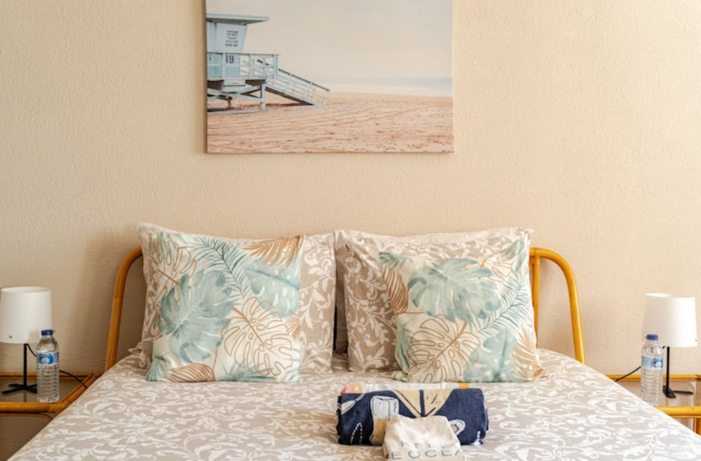 A neatly made bed with floral-patterned pillows, a water bottle, and a bag on the bed.