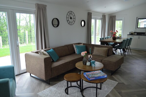 A modern living room with a sectional sofa, a dining table with chairs, a coffee table with a board game, and a kitchen area in the background. - Holiday Home Bemelen Near Nature Reserve (Bemelen)