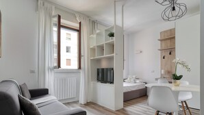A modern living room with a grey sofa, a dining area with a white table and chairs, and a built-in TV shelf.