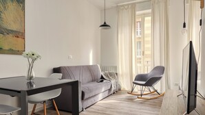 A modern living room with a grey sofa, a dining table with chairs, a rocking chair, and a flat-screen TV.