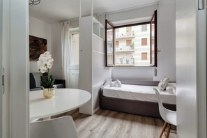 A compact room with a bed, a small table, a chair, and a window with a view of buildings. - Modern Studios near the City Centre (Milan)