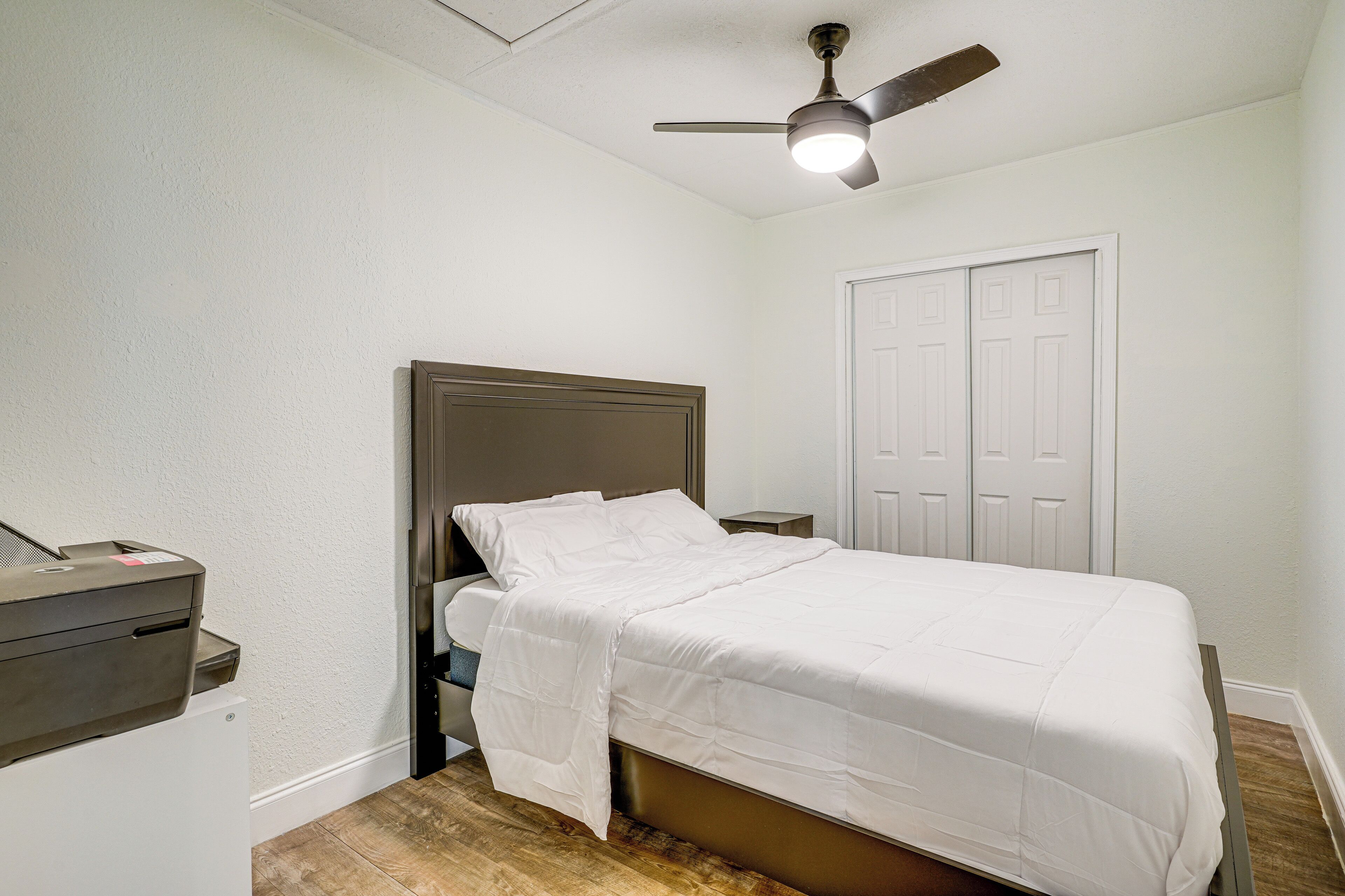 A bedroom with a bed, a ceiling fan, a white door, and a small desk with a printer.