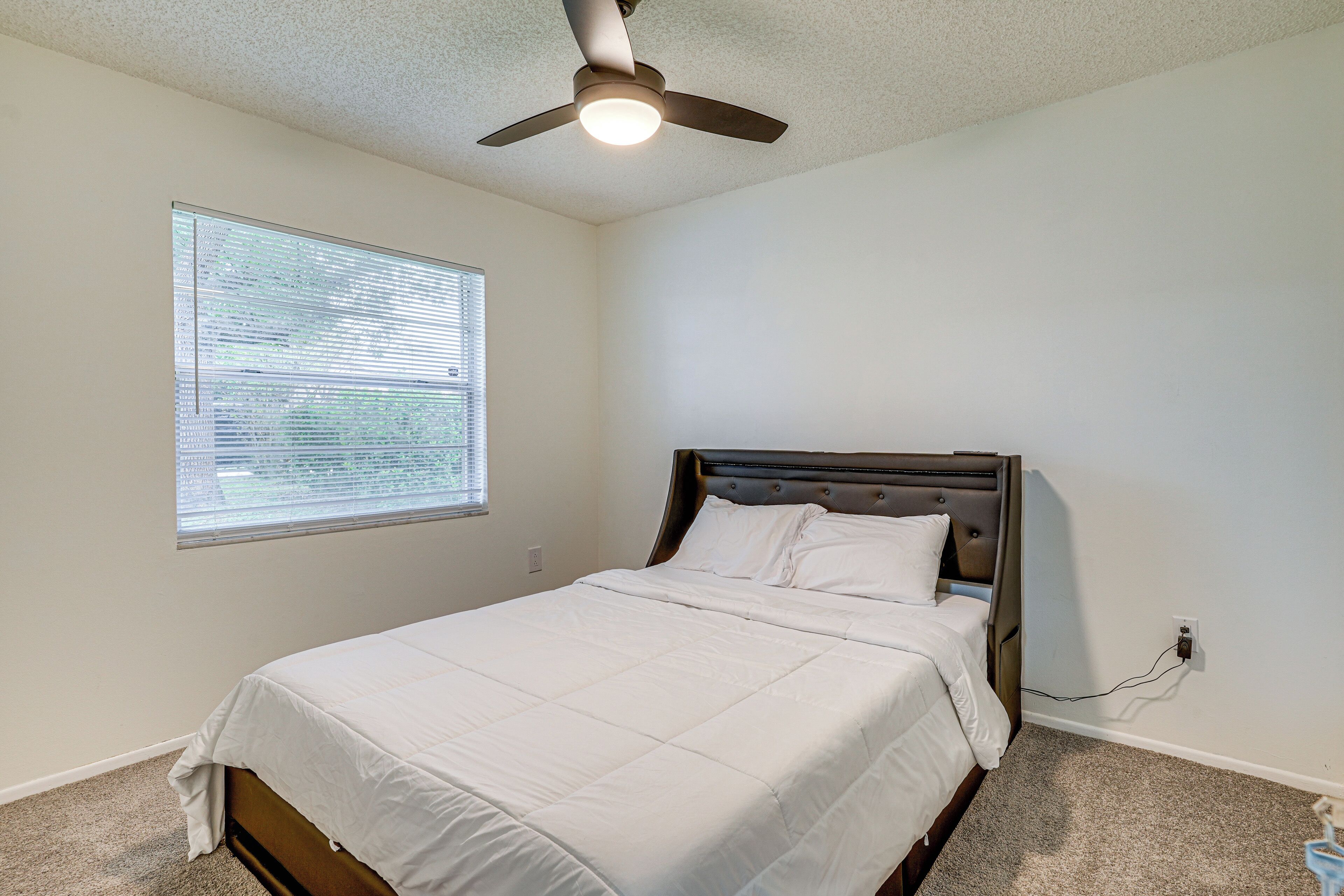 A bedroom with a bed, a ceiling fan, a window with blinds, and a power outlet on the wall.