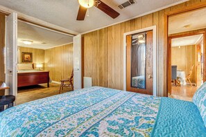 A bedroom with a wooden floor, a bed with a blue and white quilt, a wooden ceiling fan, and a wooden door leading to another room.