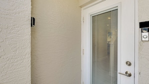 A white door with a metal handle and a keyhole, a glass panel, and a small electronic device on the wall. - Walk to Ocean: Family Retreat in Ormond Beach! (Ormond Beach)