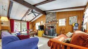 A cozy living room with a fireplace, wooden beams, a ceiling fan, and a television. - Hike, Ski & Unwind: Cabin w/ Deck in Chester! (Chester)