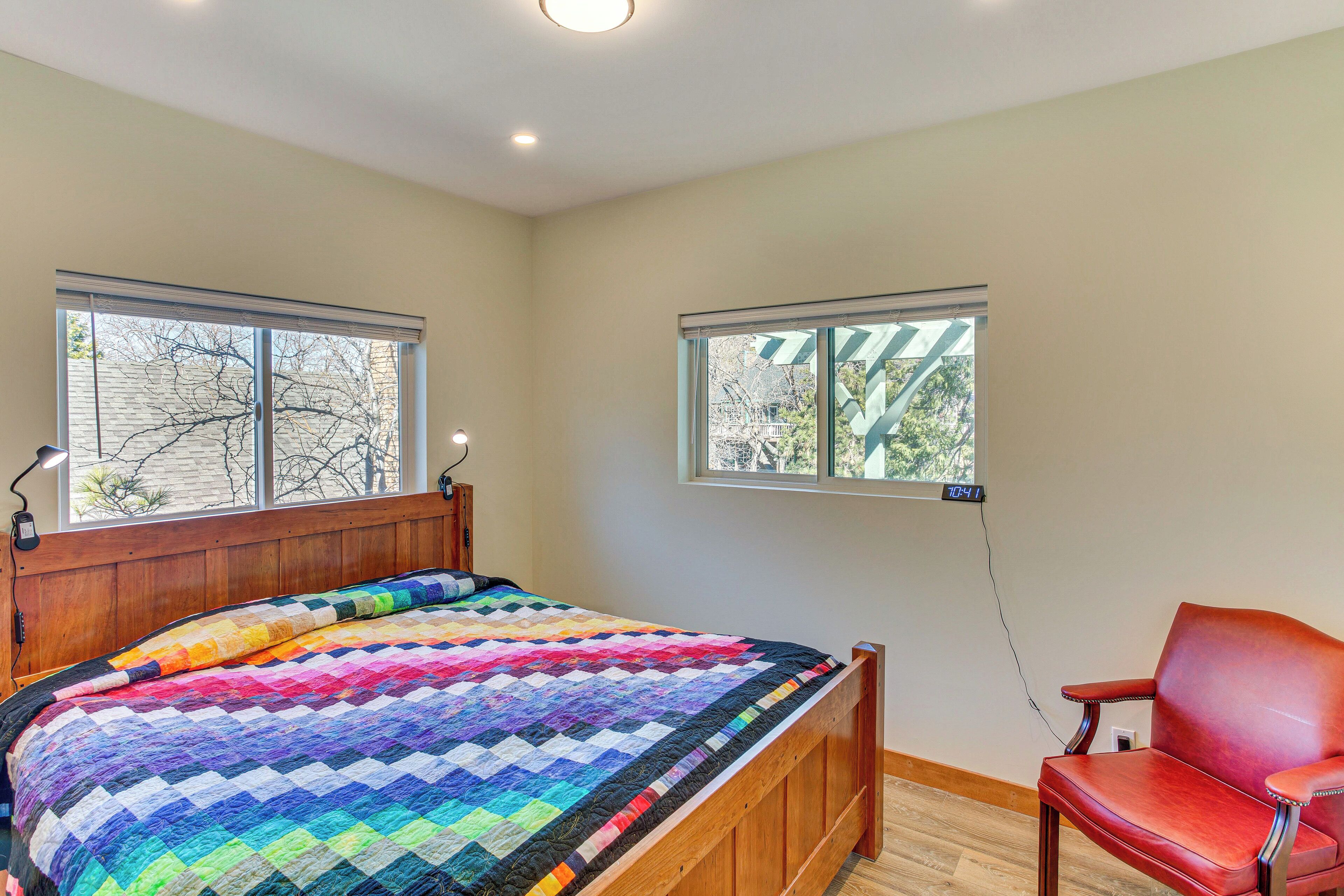 A bedroom with a wooden bed, a colorful quilt, two windows, a red chair, and a wall clock.