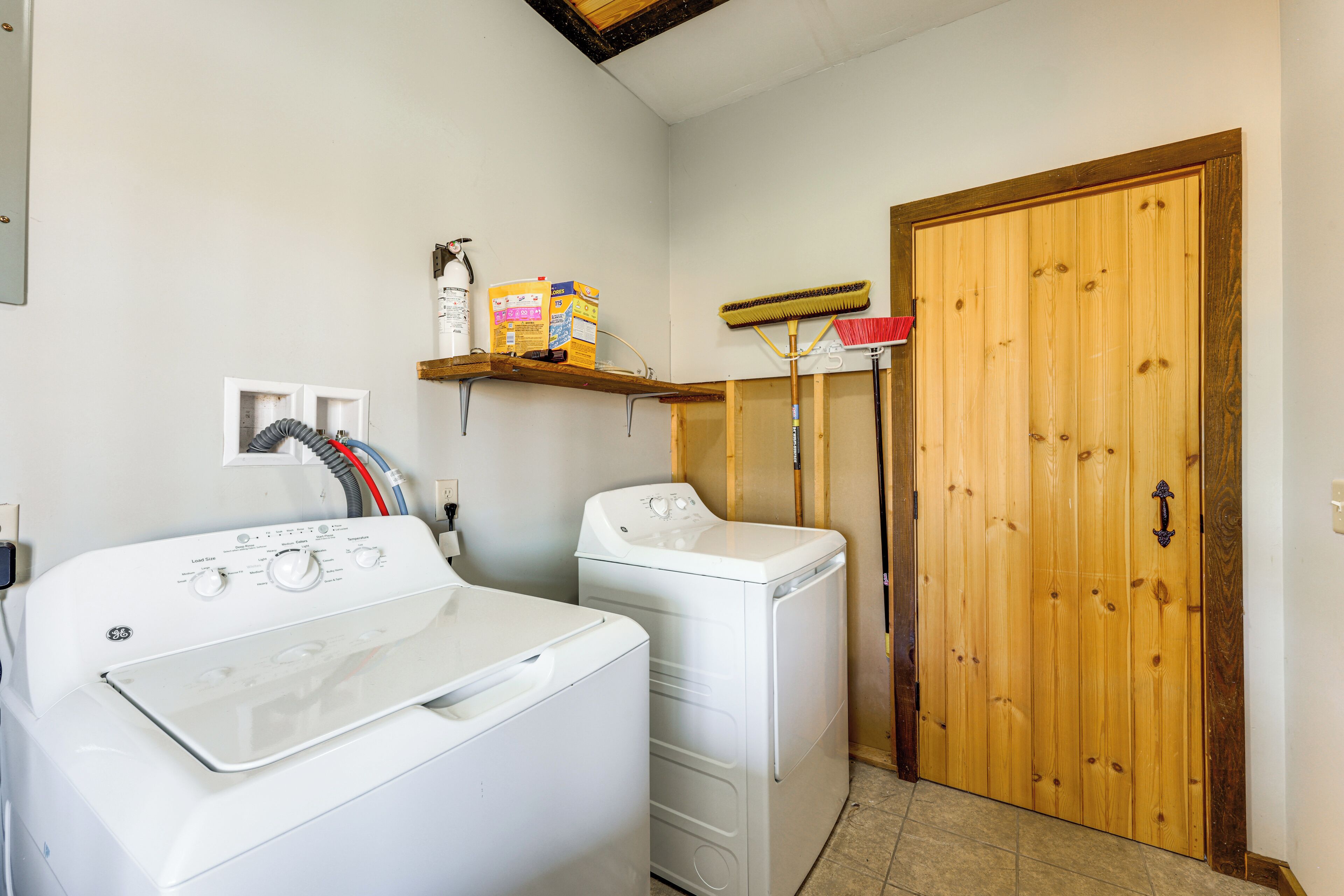 A laundry area with two white washers and dryers, a broom, and a box of detergent on a shelf.