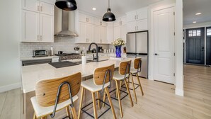 Una cocina moderna con armarios blancos, una isla de mármol, taburetes de madera e iluminación colgante.