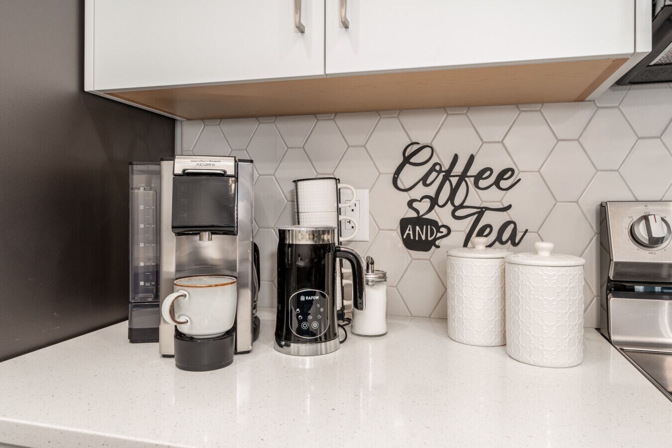 Un comptoir de cuisine avec une machine à café Hamilton Beach, une autre machine à café de la marque Rapoport, une tasse de café, un pot de sucre et un panneau indiquant « Café et thé ».