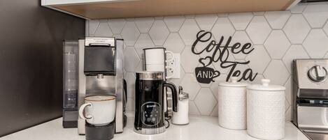 Un comptoir de cuisine avec une machine à café Hamilton Beach, une autre machine à café de la marque Rapoport, une tasse de café, un pot de sucre et un panneau indiquant « Café et thé ».