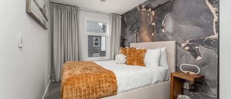 Un dormitorio moderno con una cama grande, una mesita de noche, una lámpara de pared y una ventana con cortinas.