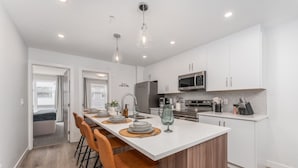 A modern kitchen with a central island, white cabinetry, stainless steel appliances, and a breakfast bar with orange chairs.