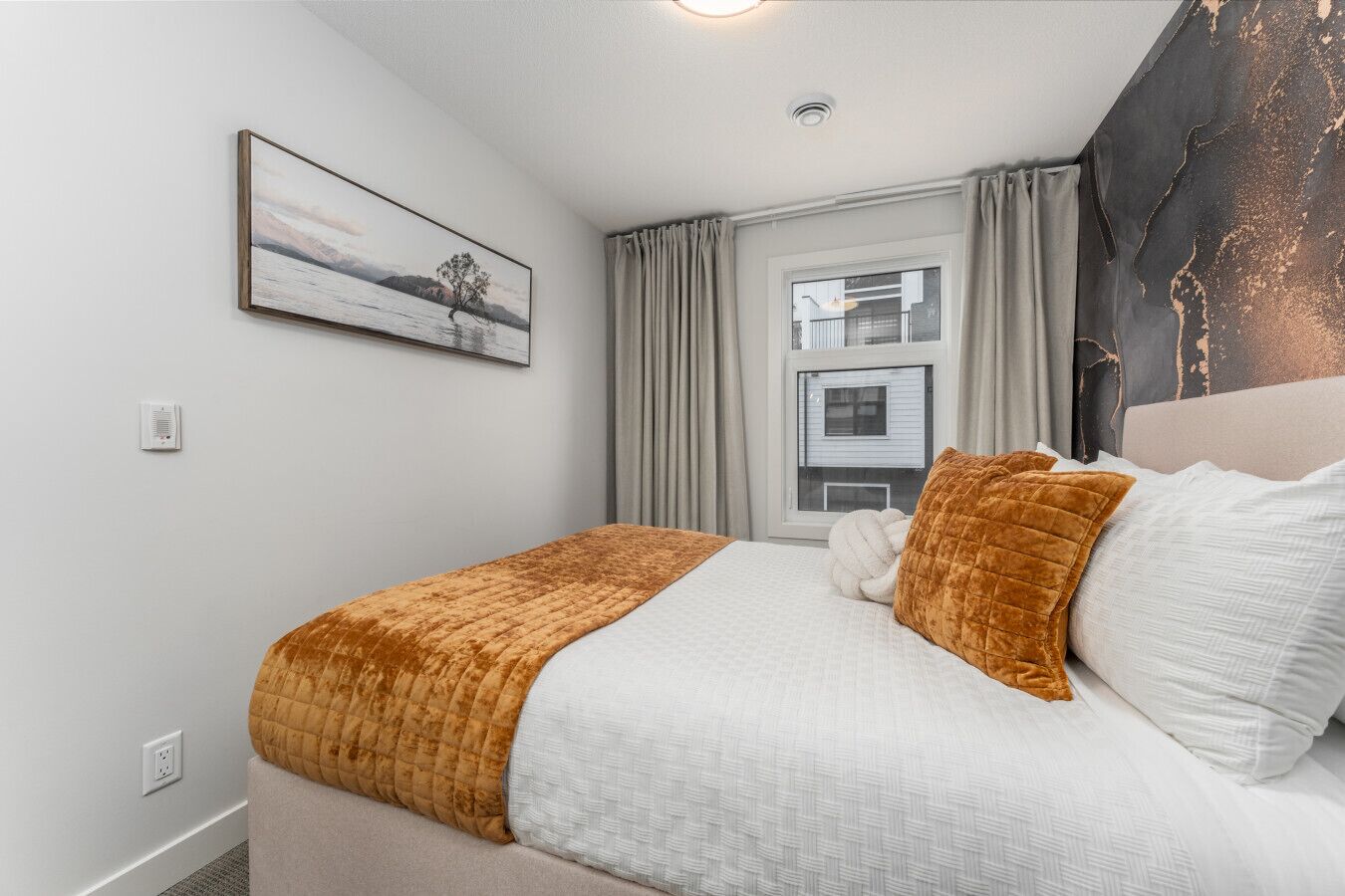 A neatly arranged hotel room with a bed, a window with curtains, a painting on the wall, and a modern ceiling light.