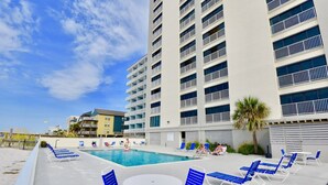 Un espace piscine avec des chaises longues bleues et une table blanche, entouré d’un grand immeuble doté de balcons.