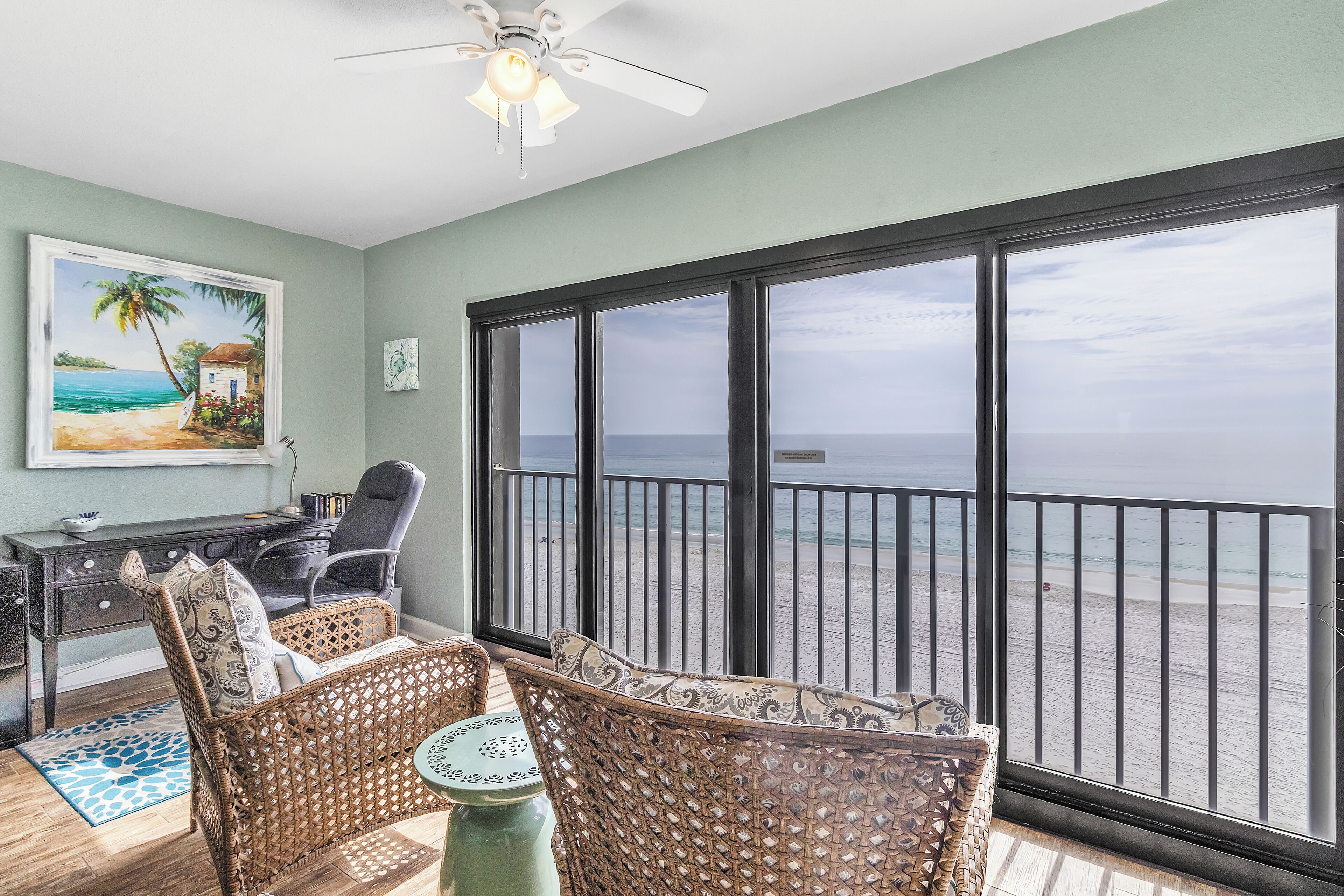A balcony with a view of the beach, wicker furniture, a desk with a chair, and a ceiling fan.