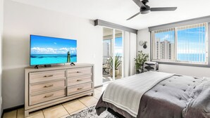 Une chambre moderne avec un lit, un téléviseur posé sur une commode, un ventilateur de plafond et un balcon offrant une vue sur la ville.