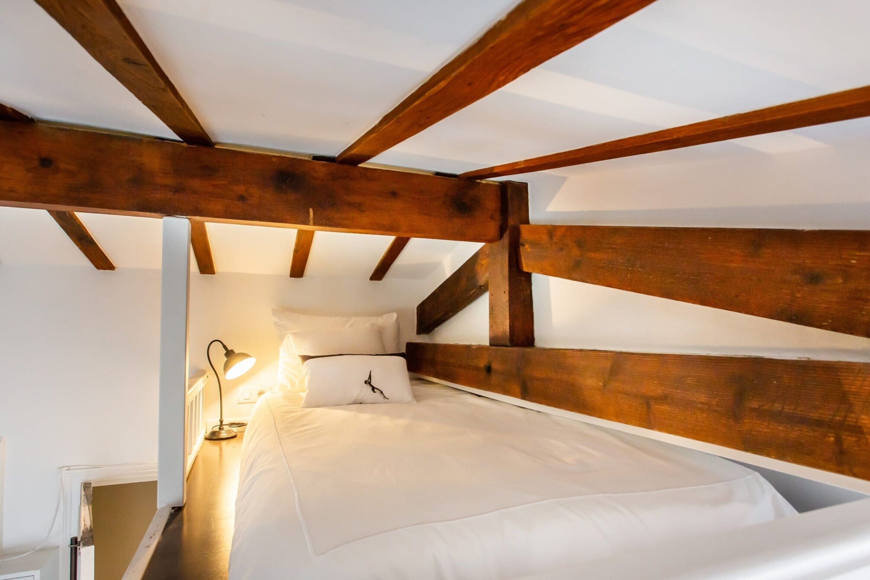 A bedroom with a wooden ceiling structure, a bed with white bedding, and a bedside lamp.