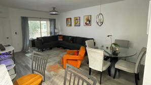 A modern living room with a sectional sofa, a dining table with chairs, and a glass coffee table. There are abstract paintings on the wall and a ceiling fan.