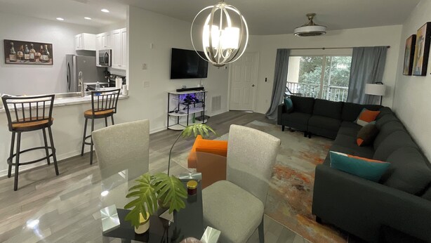 A modern living room with a dark sofa, a glass coffee table, and a dining area with a kitchen in the background.