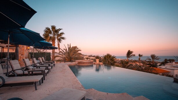 Ein Fernblick von einem Infinity-Pool mit Liegestühlen und Sonnenschirmen auf eine Küstenlandschaft mit Palmen und einem Sonnenuntergang.