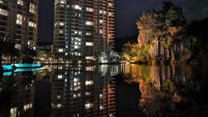A high-rise building with multiple floors, a reflective water body in front, and a rocky outcrop with trees. - Mountain & Lake Views' Suite Cycas (Ipoh)