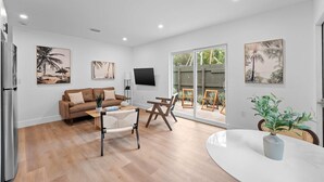Una sala moderna con un sofá, una mesa de centro y una mesa de comedor con sillas. Hay pinturas en las paredes y un televisor montado sobre el sofá.