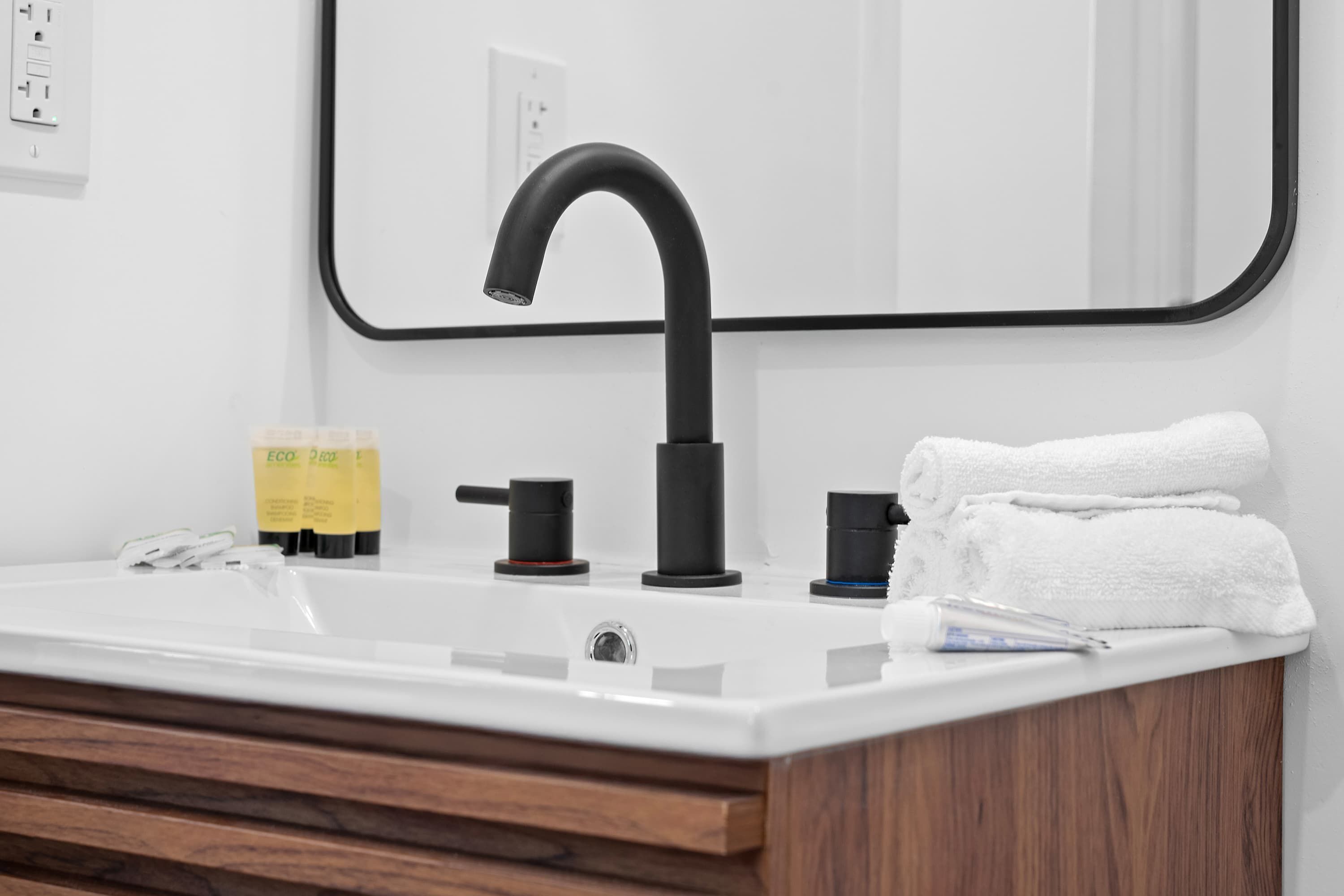 Een badkamer met een witte wastafel, een zwarte kraan en een houten kastje. Er staan toiletartikelen op het aanrecht en een handdoek is rechts gestapeld.