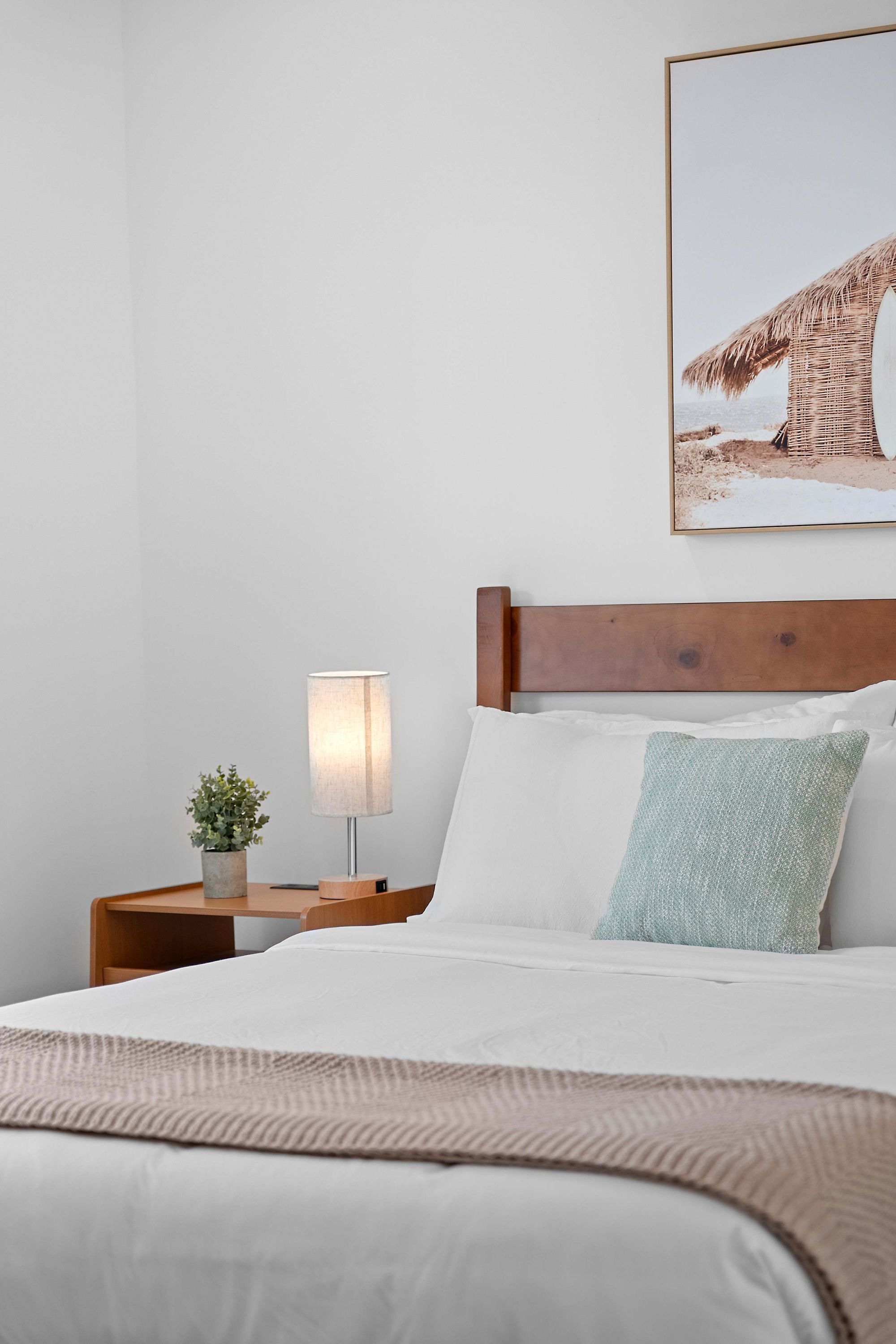 Un letto rifatto con cura, una testiera in legno, una lampada da comodino, una piccola pianta e un quadro appeso al muro.