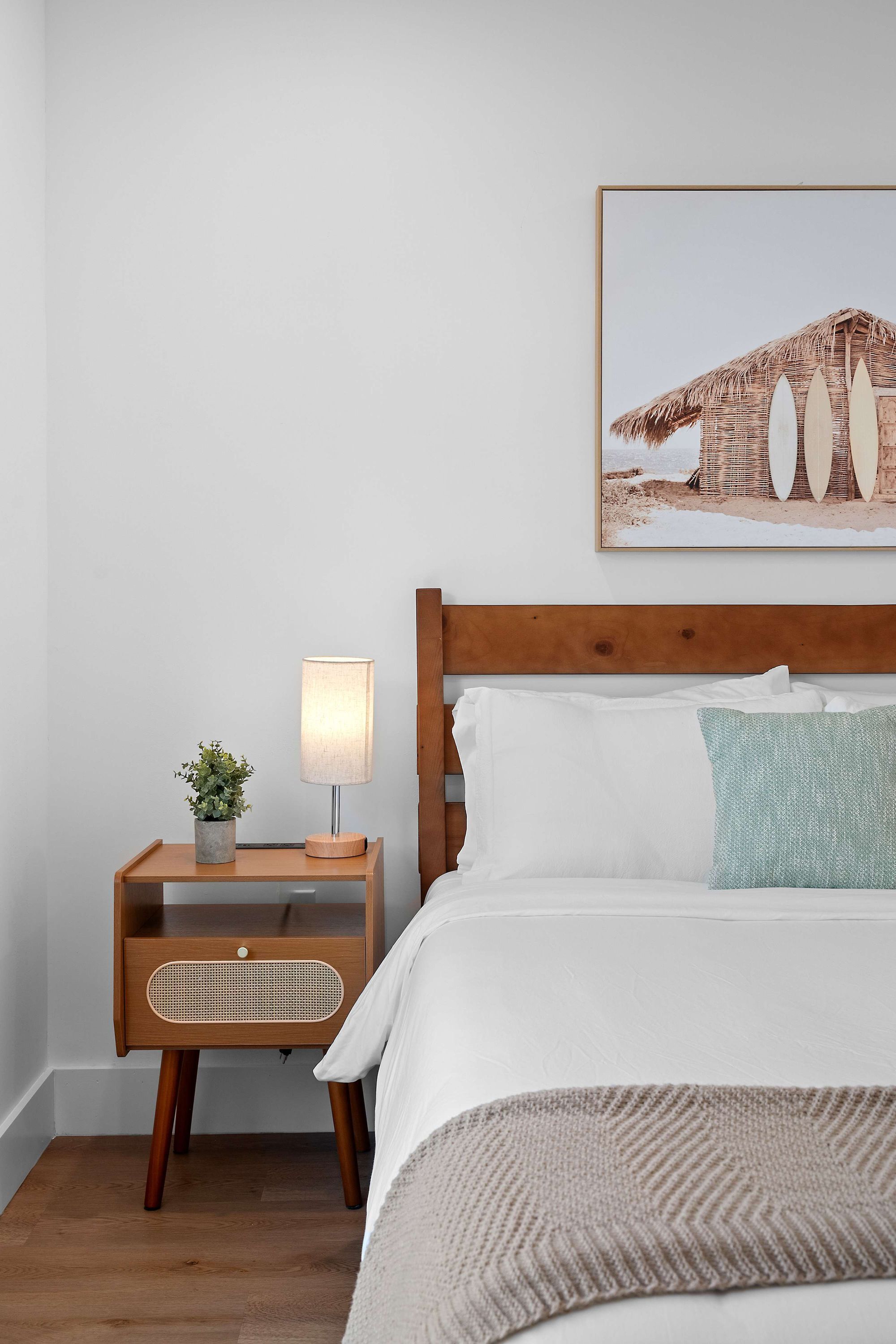 A neatly arranged bedroom with a wooden headboard, a bedside table with a lamp, a small plant, and a framed picture on the wall.