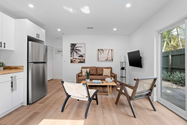 A modern kitchen with white cabinets, a stainless steel refrigerator, and a wooden countertop. There is a brown sofa with a plant and a coffee table in the living area. - 921 Apartment 1 in Fort Lauderdale (Fort Lauderdale)