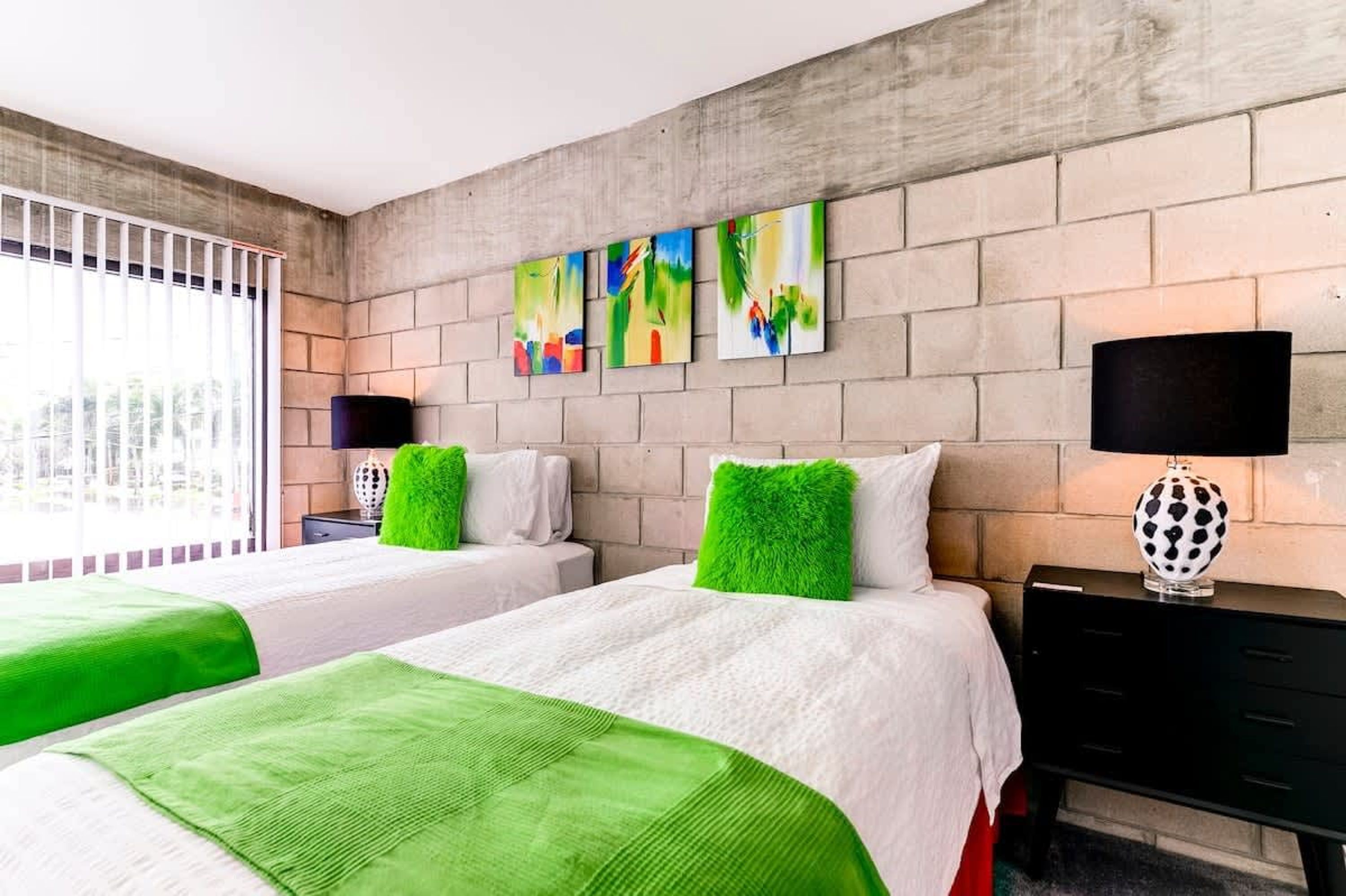 Quarto de hotel com duas camas, roupa de cama verde e branca, parede de tijolos e obras de arte sobre as camas.