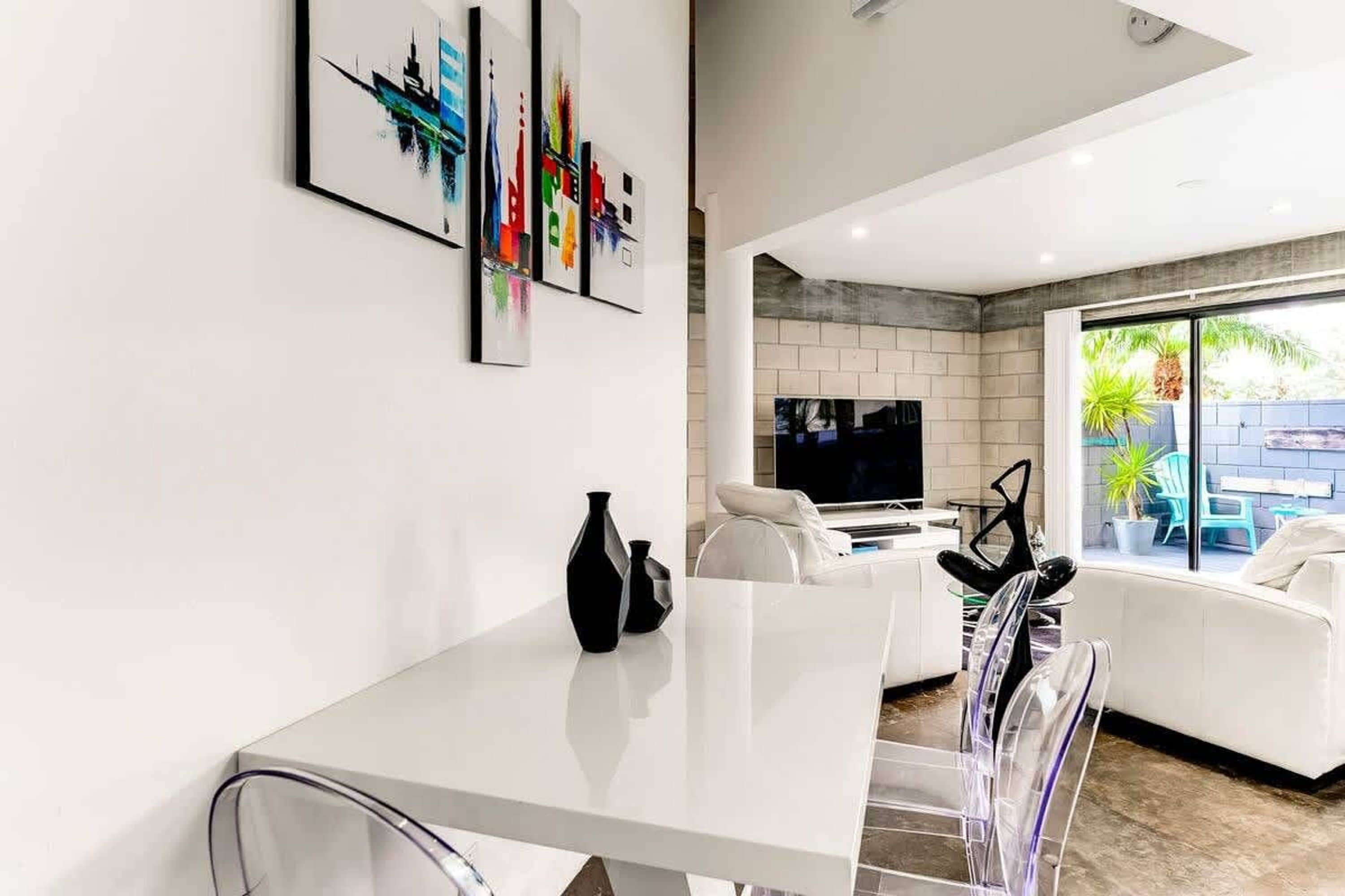Uma sala de estar moderna com um sofá branco, uma mesa de centro de vidro e uma TV. Há quadros abstratos na parede e uma janela com vista para o exterior.