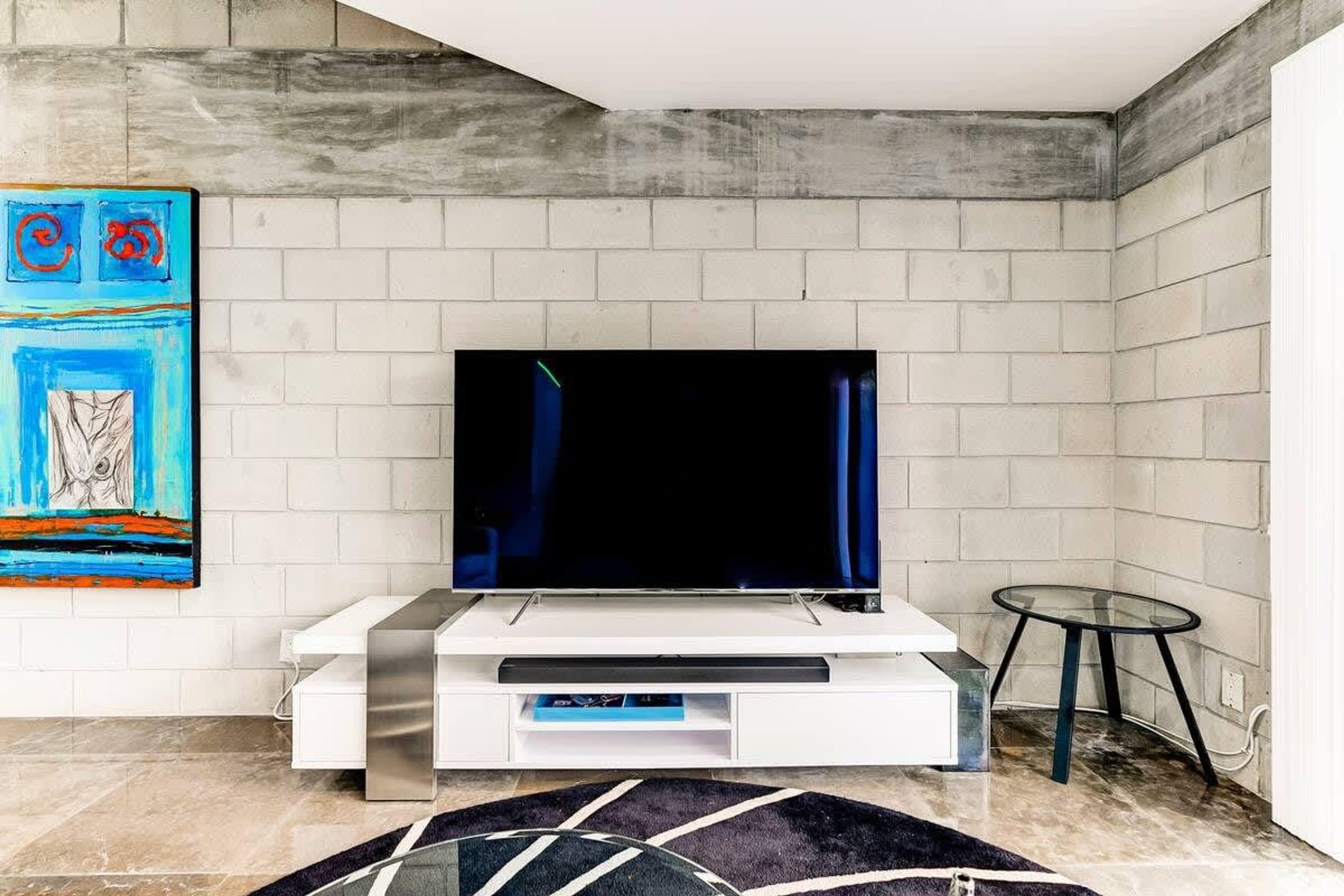 Un salon moderne avec un téléviseur à écran plat posé sur un meuble de télévision blanc, une table basse en verre et une peinture abstraite aux couleurs vives accrochée au mur.