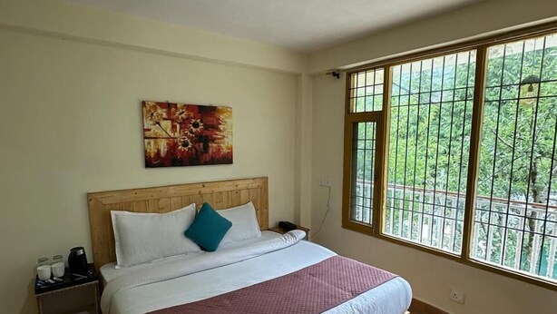 A bedroom with a wooden bed, a nightstand, a painting, and a window with a view of greenery. - The Nature Crest Manali (Manali)