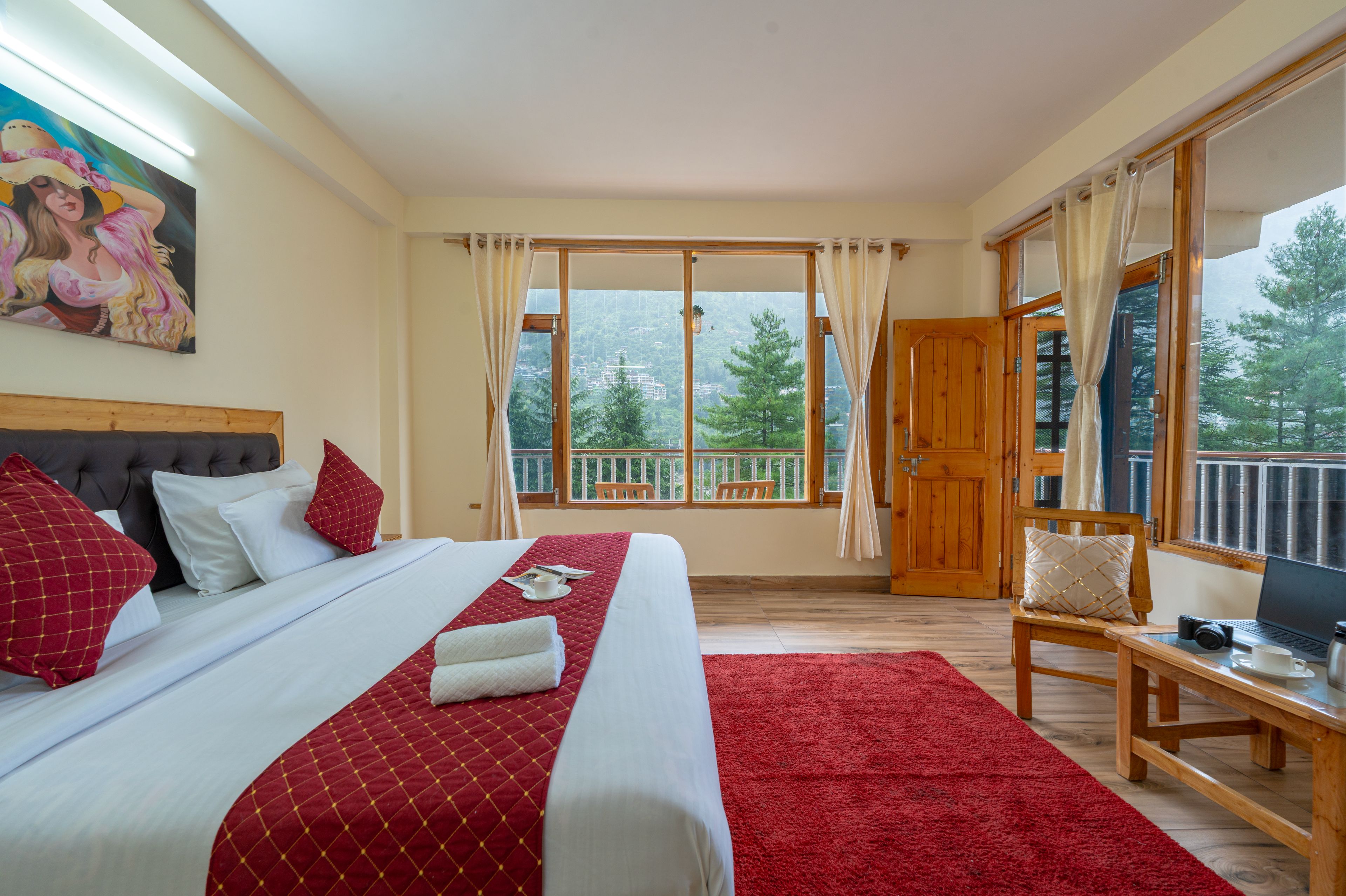 A hotel room with a large bed, a desk with a laptop, a chair, and a balcony with a view of trees and buildings.
