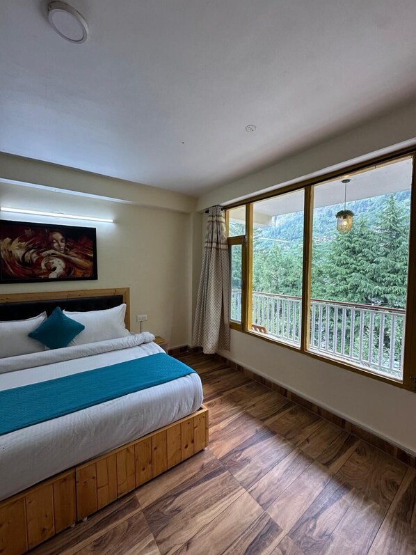 A bedroom with a large window, wooden flooring, a bed with white bedding, and a painting on the wall. - The Nature Crest Manali (Manali)