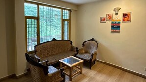 A room with a sofa, armchair, and coffee table, large windows with a view of trees, and wall decorations. - The Nature Crest Manali (Manali)