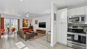 Una sala de estar moderna con sofá, mesa de centro y televisor, contigua a una cocina con armarios blancos y electrodomésticos de acero inoxidable.