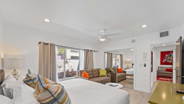 Habitación de hotel con una cama grande, un sofá, una mesita de café y un televisor. Hay cortinas en las ventanas y un ventilador de techo.