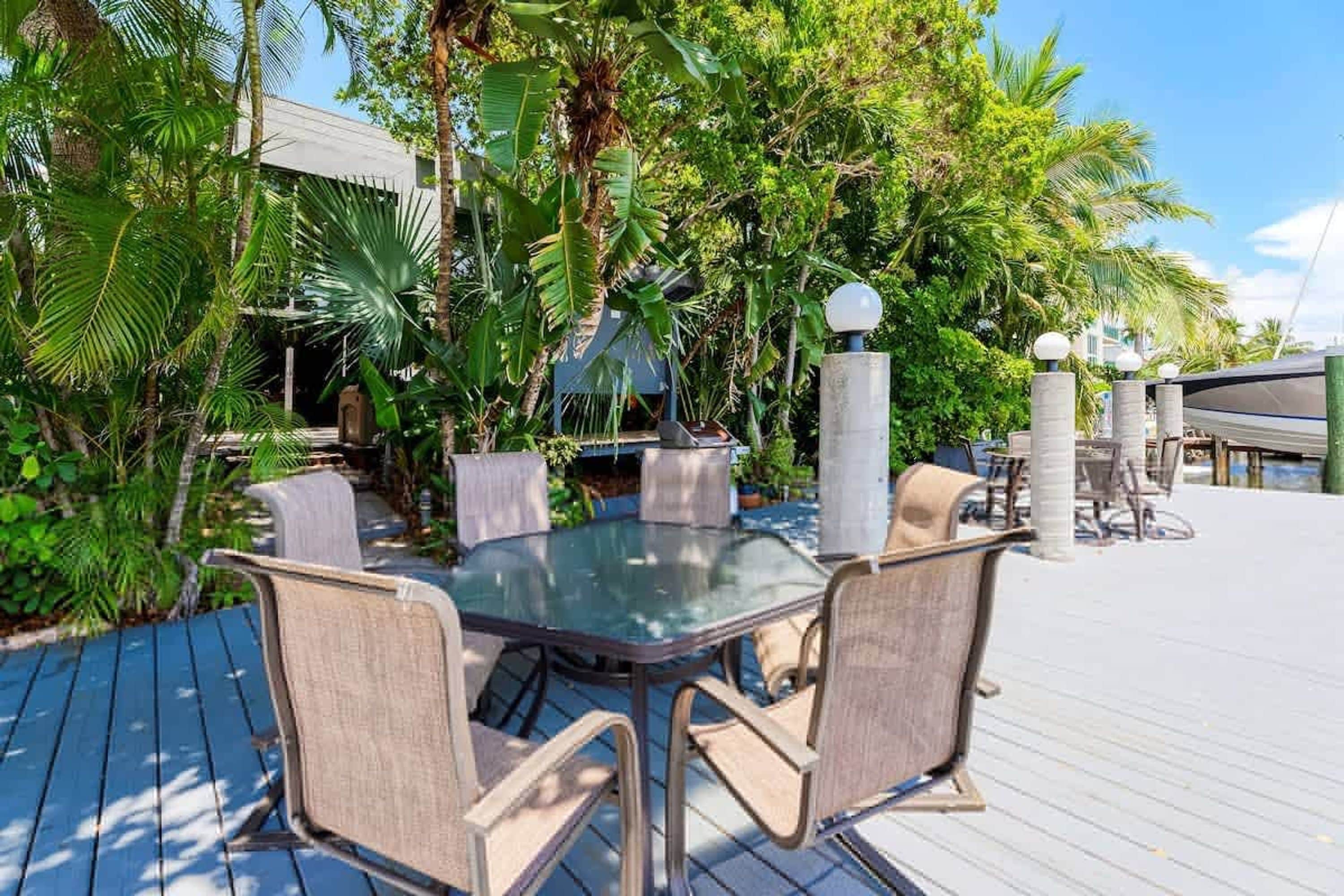 An outdoor dining area with a glass table and cushioned chairs, surrounded by lush tropical plants and a wooden deck.