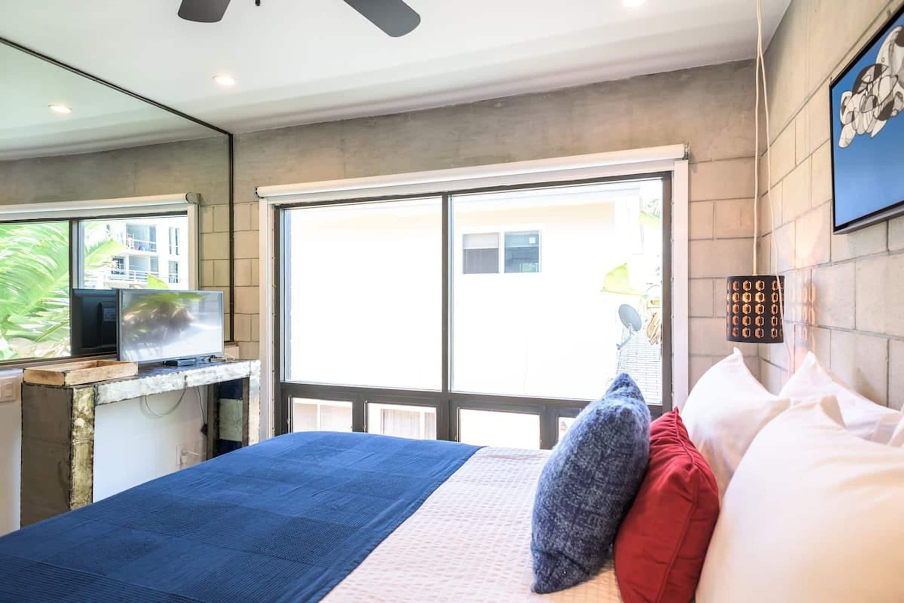 Une chambre d’hôtel moderne avec un grand lit, un téléviseur posé sur un meuble en bois, une grande fenêtre et un ventilateur de plafond.