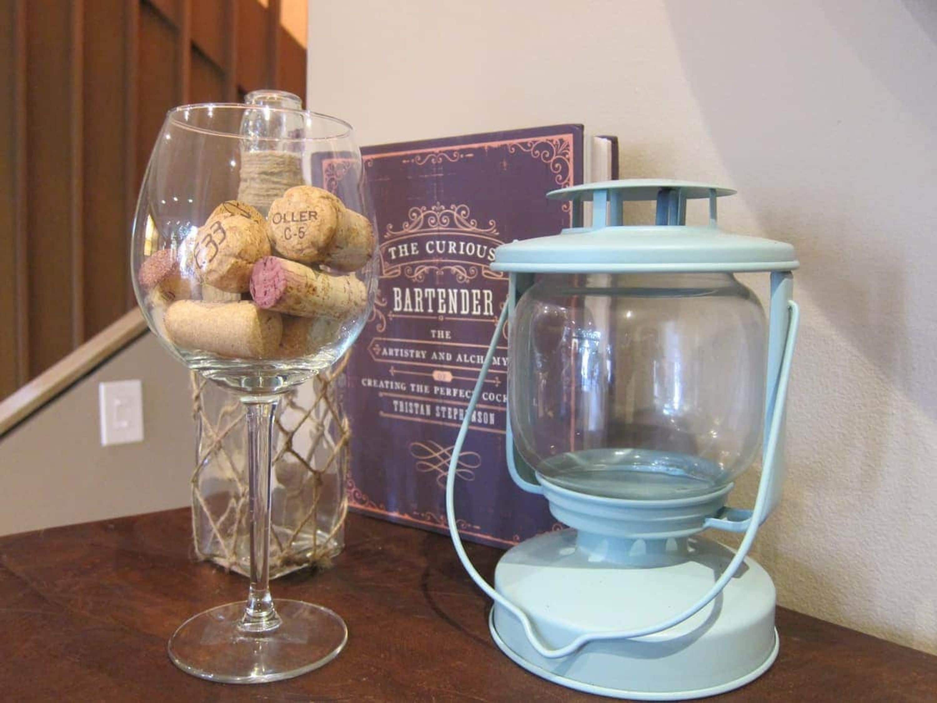 Un florero de vidrio con corchos, un libro titulado 'The Curious Bartender' y un farol azul sobre una mesa de madera.