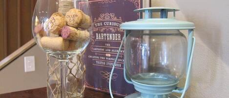 Un florero de vidrio con corchos, un libro titulado 'The Curious Bartender' y un farol azul sobre una mesa de madera.