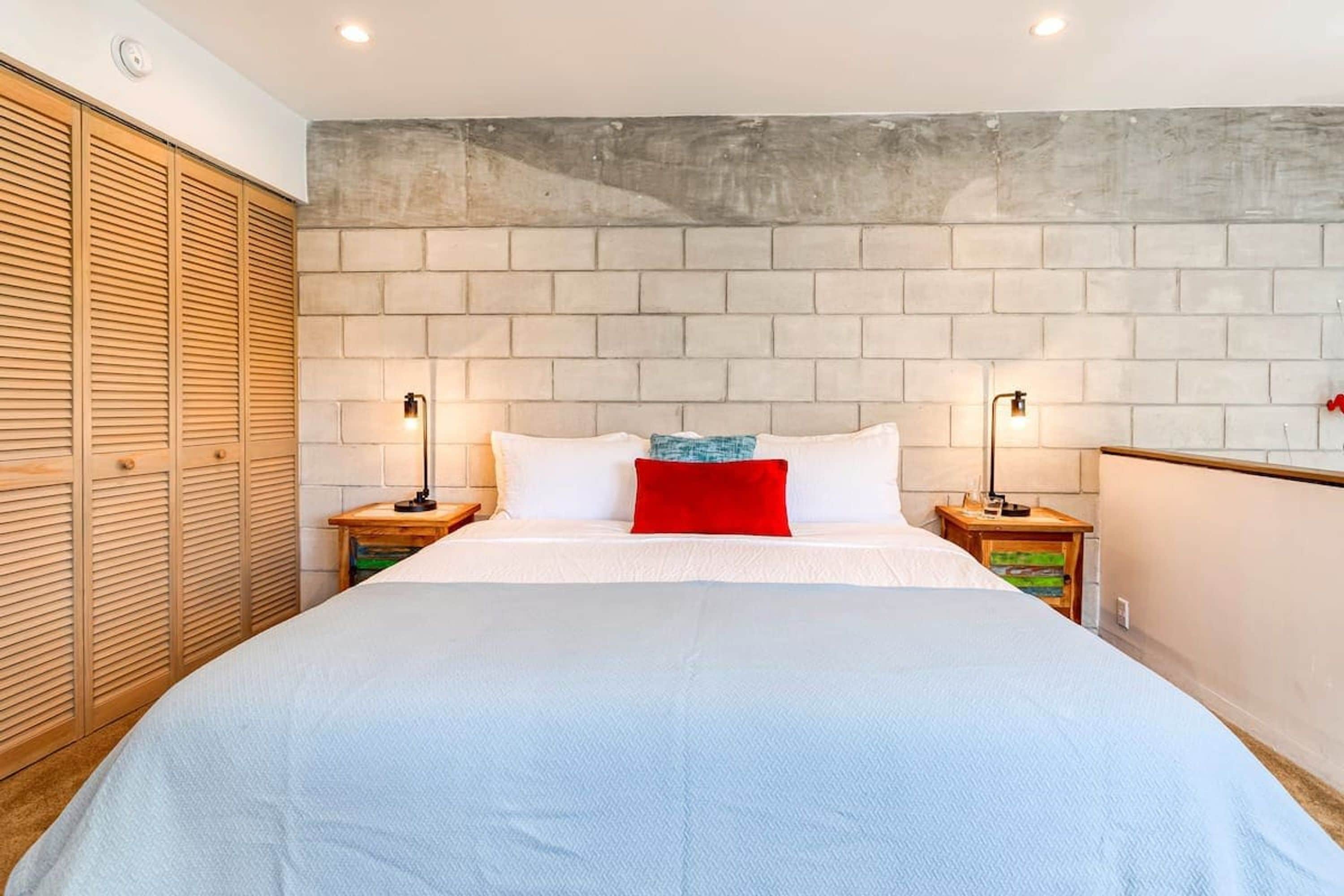 A bedroom with a large bed, two bedside tables with lamps, a wooden wardrobe, and a brick-patterned headboard.