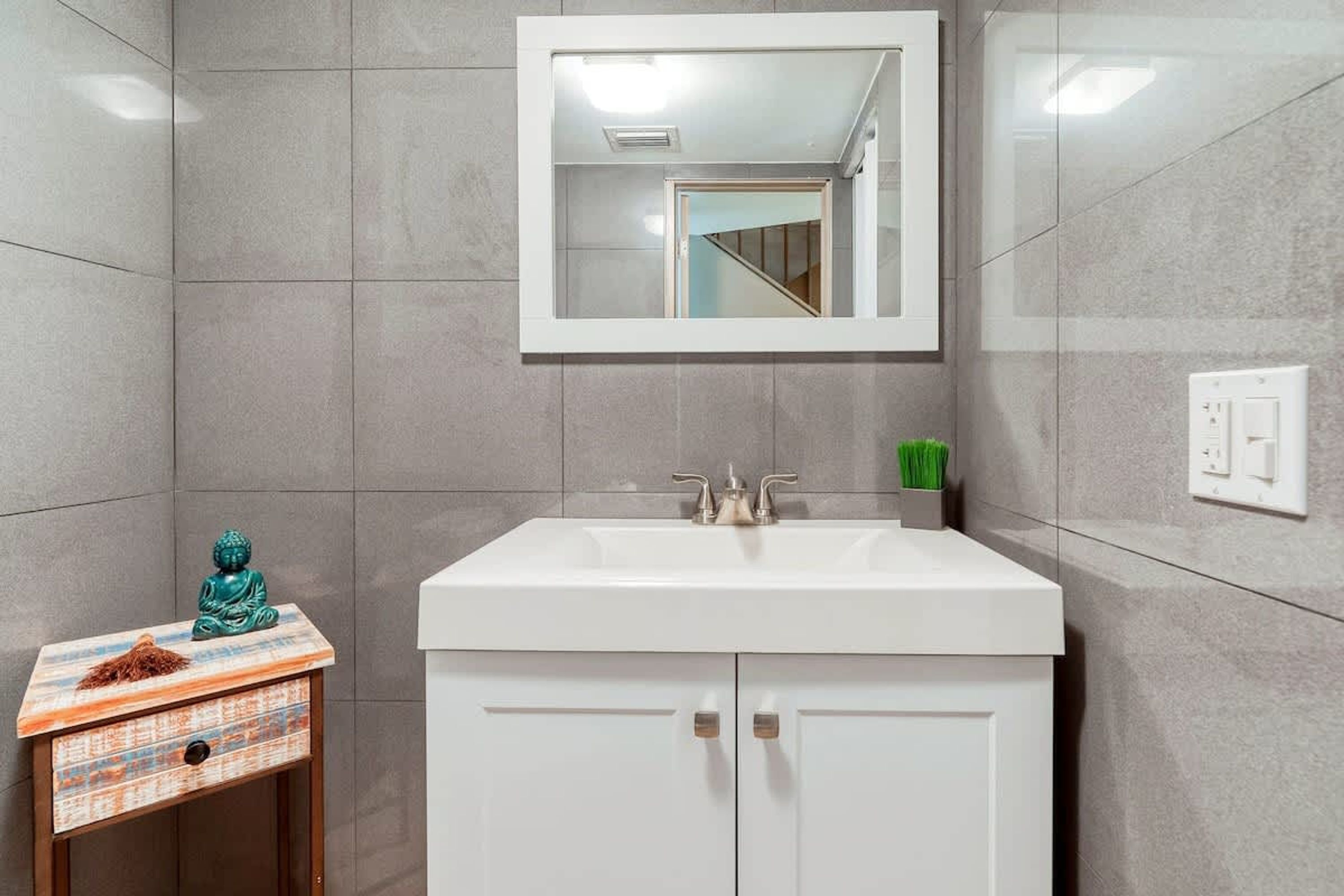 A bathroom with a white sink, a small wooden side table with a decorative Buddha, and a mirror above the sink.