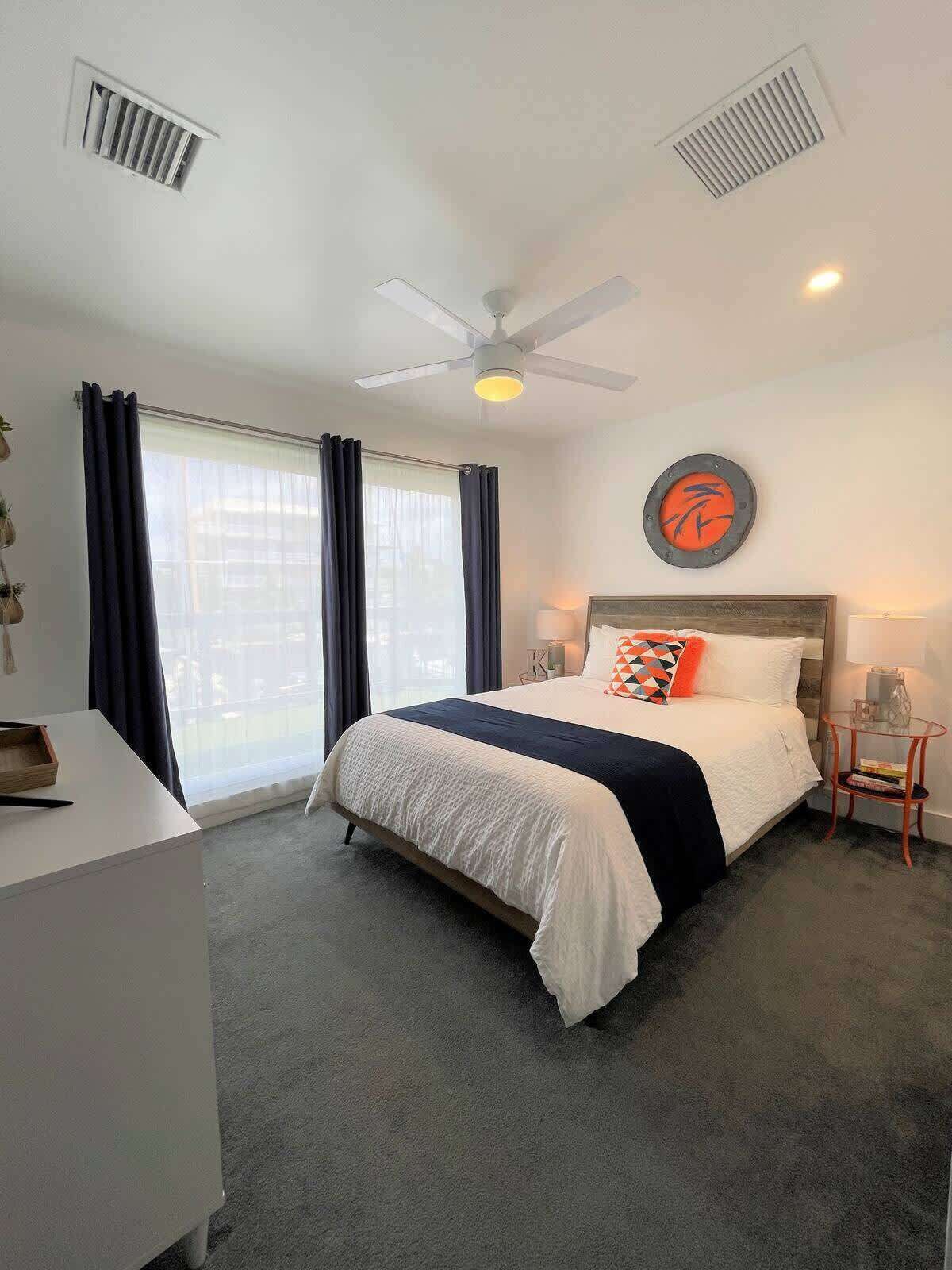 Une chambre d’hôtel équipée d’un lit, d’un bureau, d’une chaise, d’un ventilateur de plafond et d’une fenêtre avec des rideaux.
